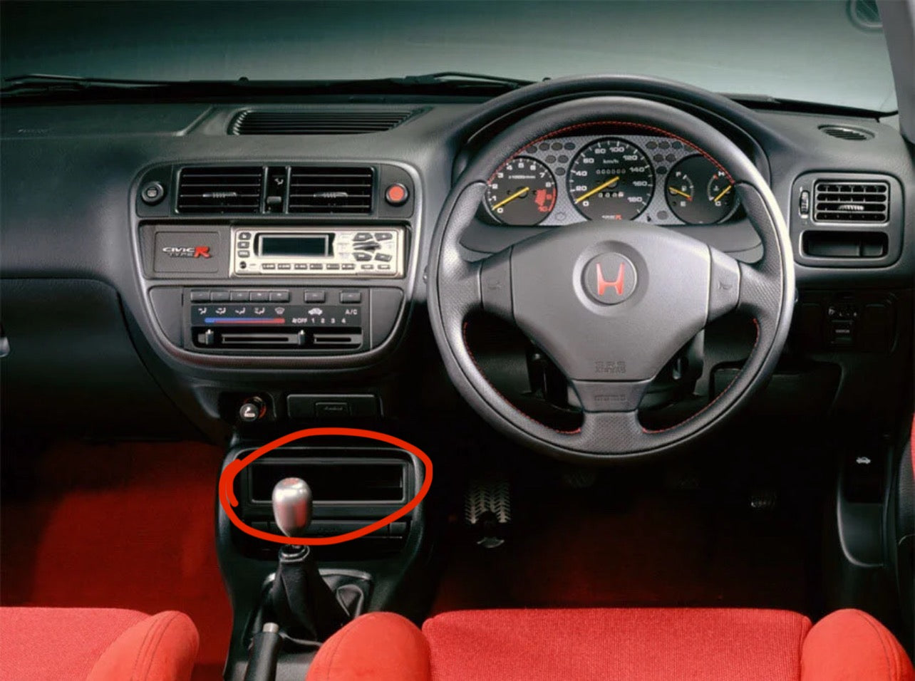 A close-up view of a car interior showing the center console with a manual gear shift stick and a red handbrake, highlighted by a red circle. The dashboard, steering wheel with a Honda logo, and red seats are visible.