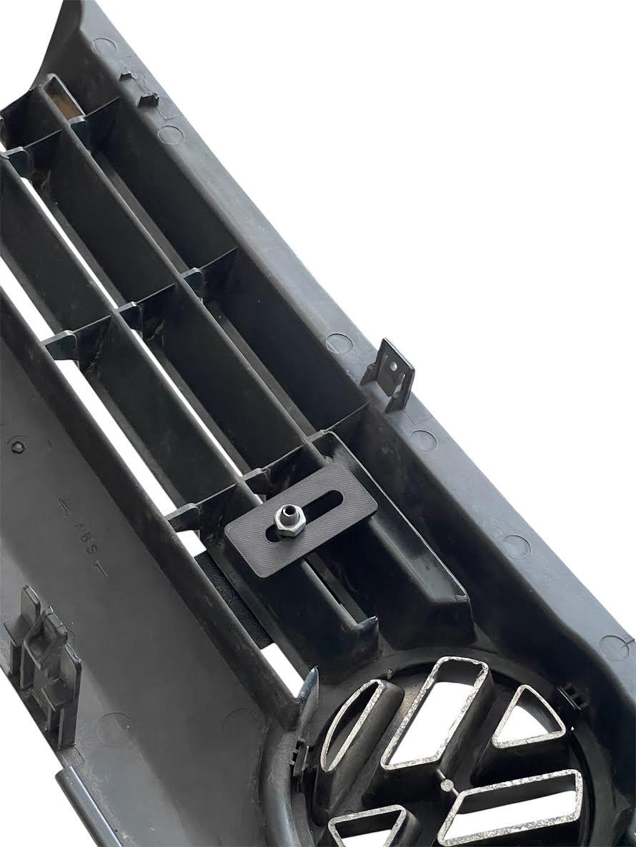 Rear view of the Volkswagen Motorsport emblem attached to a VW front grille, showing the mounting bracket and screw installation method.