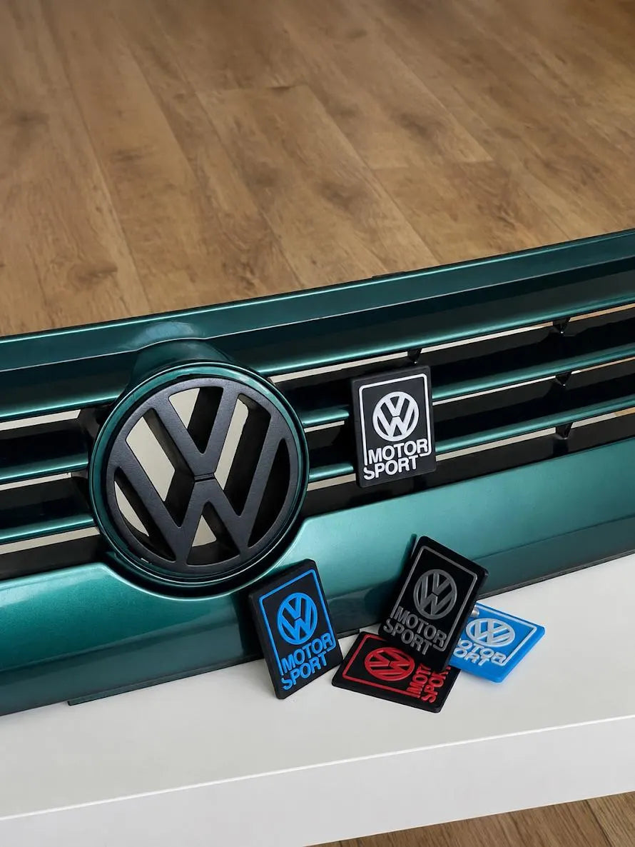 Collection of 3D printed Volkswagen Motorsport emblems installed on a Volkswagen front grille, showing all available mounting and design options.